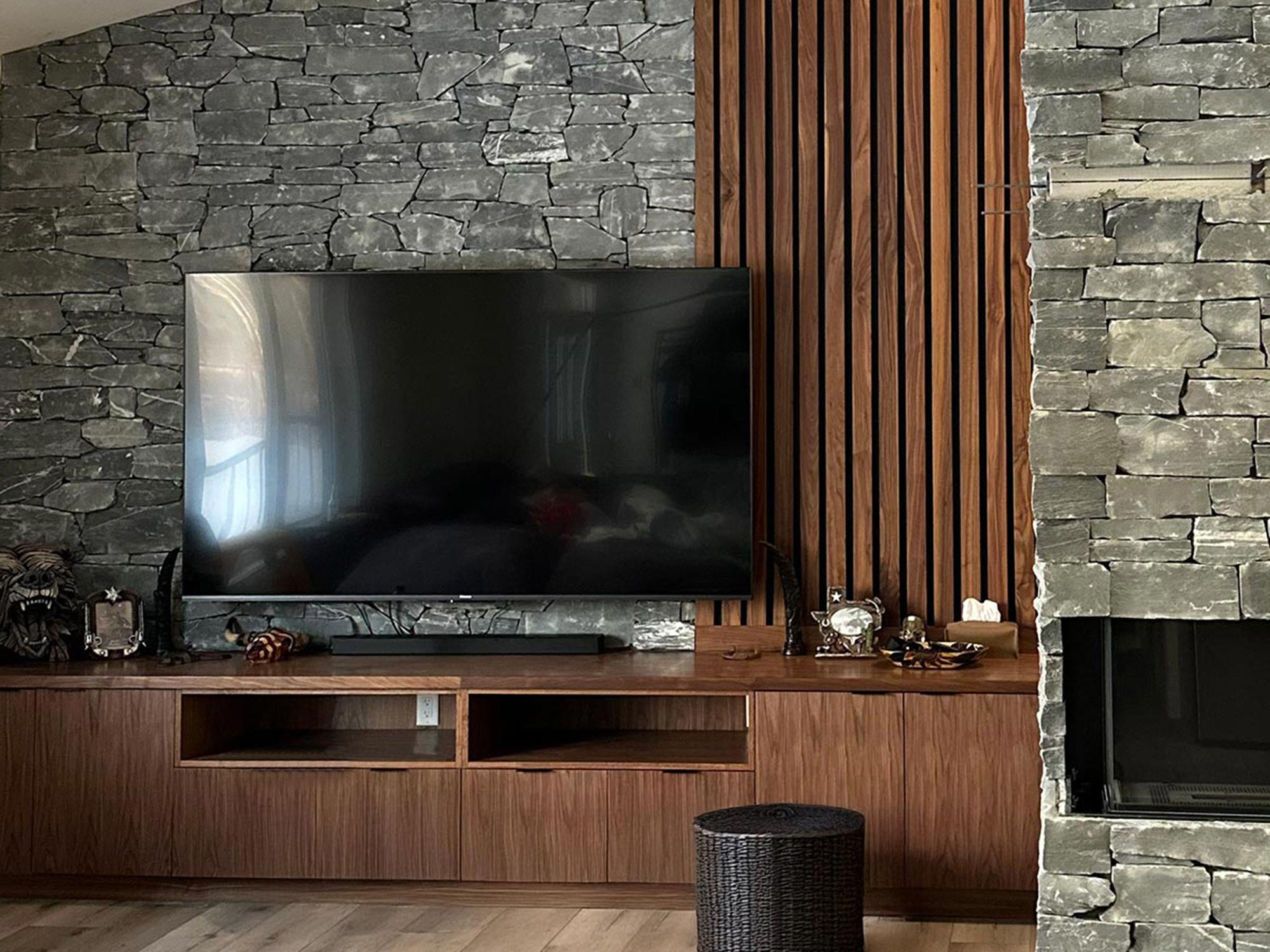 Dark millwork / woodwork television console stretches from wall to wall, with vertical wood strips forming a panel from the top of the console to the ceiling along the right side of a grey stonework wall. The wood panel/feature follows the angled rise of the ceiling. A stone-faced gas fireplace sits to the right in the image.