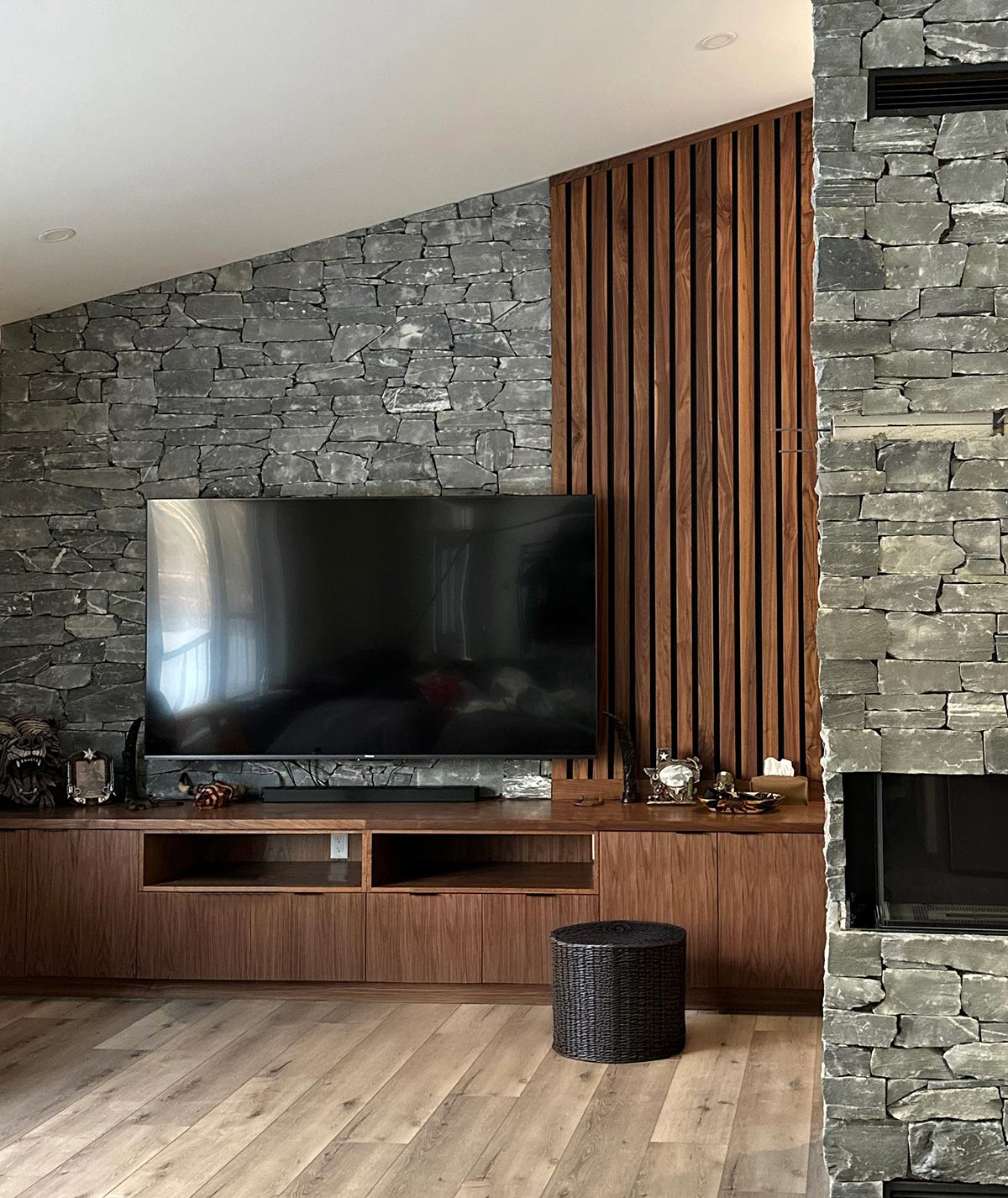Dark millwork / woodwork television console stretches from wall to wall, with vertical wood strips forming a panel from the top of the console to the ceiling along the right side of a grey stonework wall. The wood panel/feature follows the angled rise of the ceiling. A stone-faced gas fireplace sits to the right in the image.